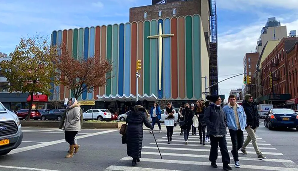 Pedestrians are crossing a street in an urban setting with a distinctive building adorned with a large cross in the background