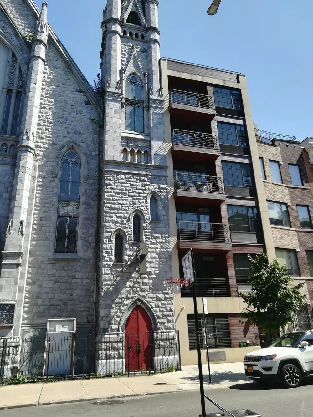 The image shows a contrast between a traditional stone church with a red door and a modern residential building with balconies under a clear blue sky