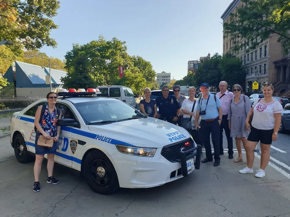 A group of people pose for a photo with two police officers next to an NYPD police car on a sunny day
