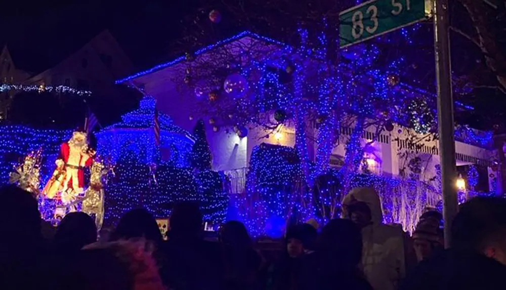A crowd of people admires an extravagant holiday light display featuring a large illuminated Santa Claus figure and a house covered in blue lights at night