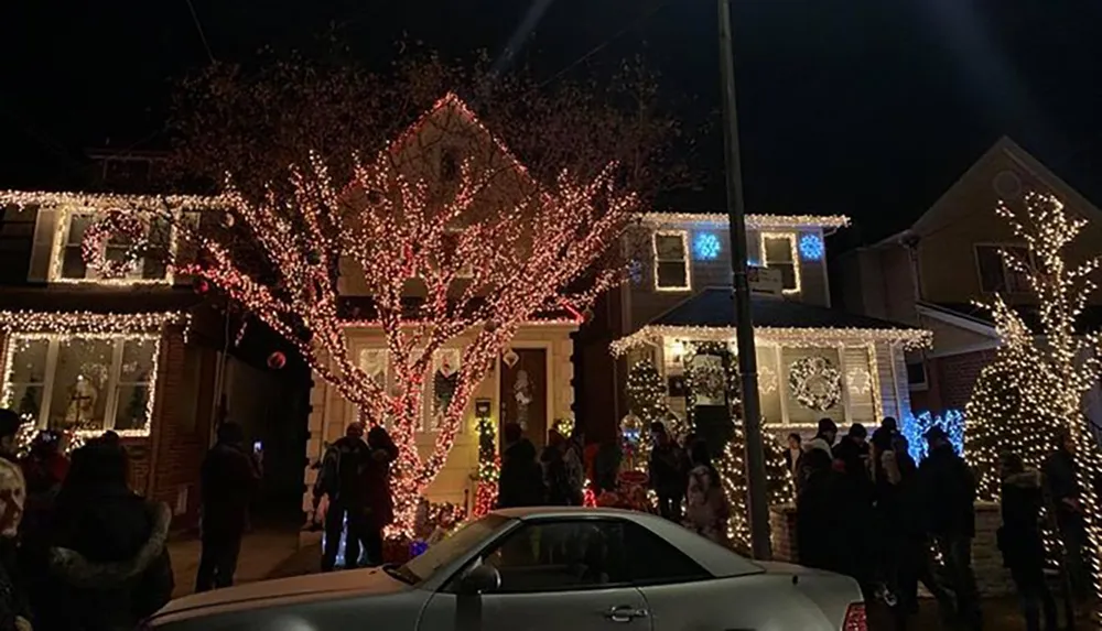 The image shows a festive scene where houses and trees are adorned with colorful Christmas lights with people gathered on the street to enjoy the decorations