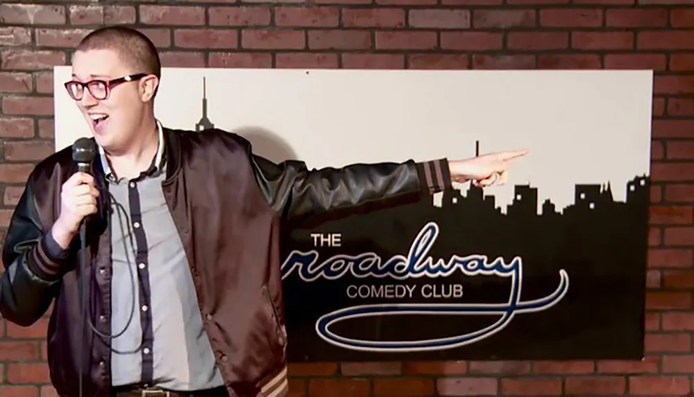 A comedian is performing on stage at The Broadway Comedy Club enthusiastically pointing to something while holding a microphone