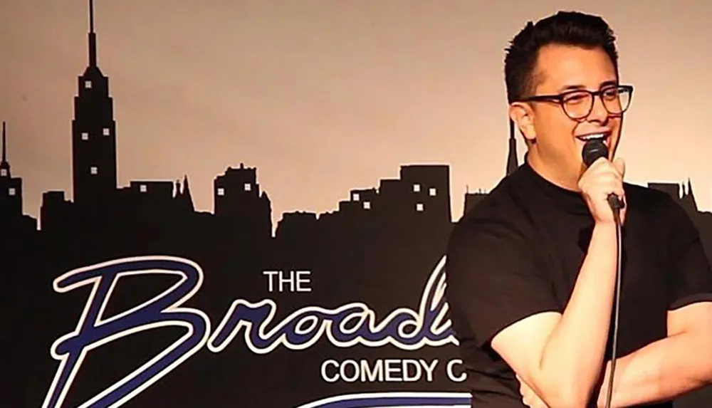 A person is performing on stage at a comedy club with a microphone in hand and a backdrop featuring the silhouette of a city skyline and the text The Broadway Comedy Club