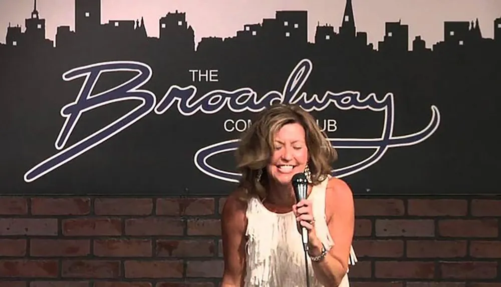 A person is performing on stage at The Broadway Comedy Club as indicated by the backdrop