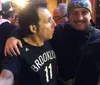 Two people are smiling for a photo one in a Brooklyn basketball jersey and the other giving a thumbs up with someone else taking a photo of them in the foreground