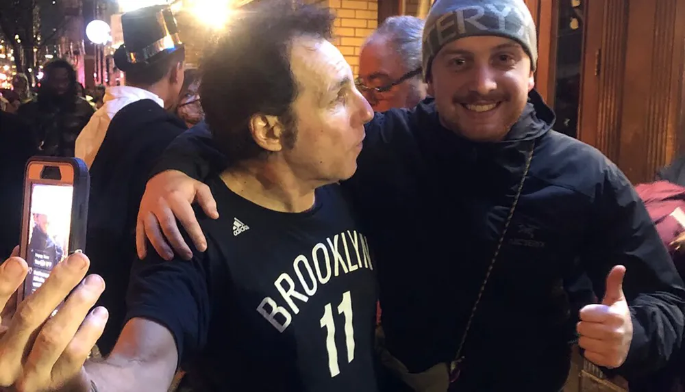 Two people are smiling for a photo one in a Brooklyn basketball jersey and the other giving a thumbs up with someone else taking a photo of them in the foreground