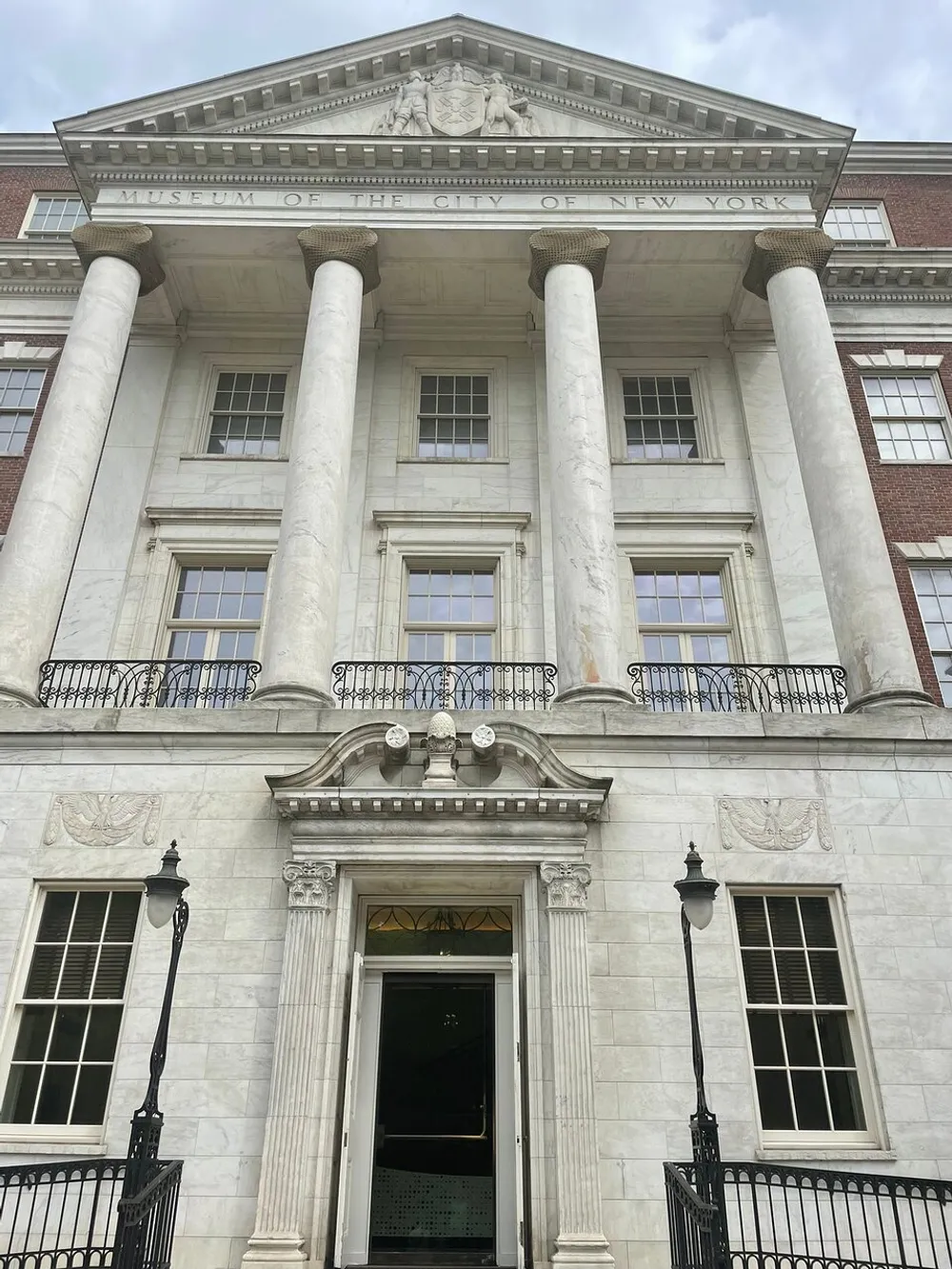 The image shows the neoclassical facade of the Museum of the City of New York with its grand entrance flanked by towering columns and inscription above