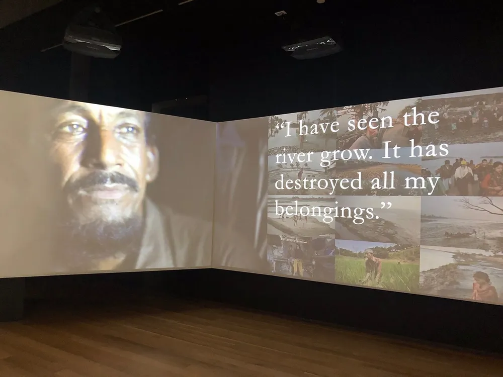 The image shows a multimedia installation with a close-up portrait of a man projected onto a screen alongside panels with the quote I have seen the river grow It has destroyed all my belongings and smaller accompanying photographs presumably related to the rivers impact