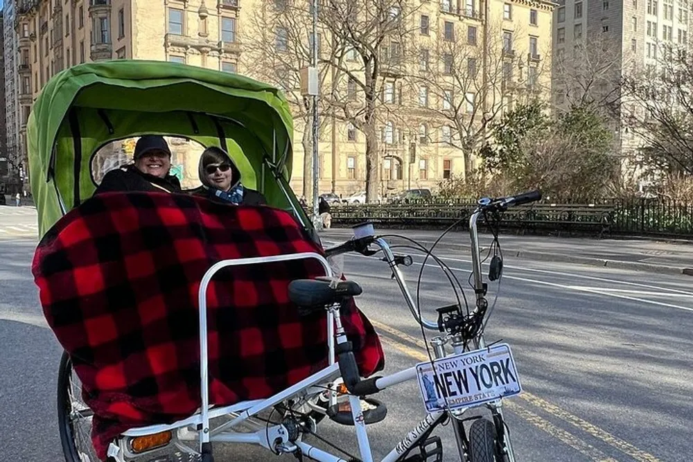 Two people are sitting under a red and black blanket in a green pedicab labeled New York on a street that appears to be in an urban area with trees and buildings in the background