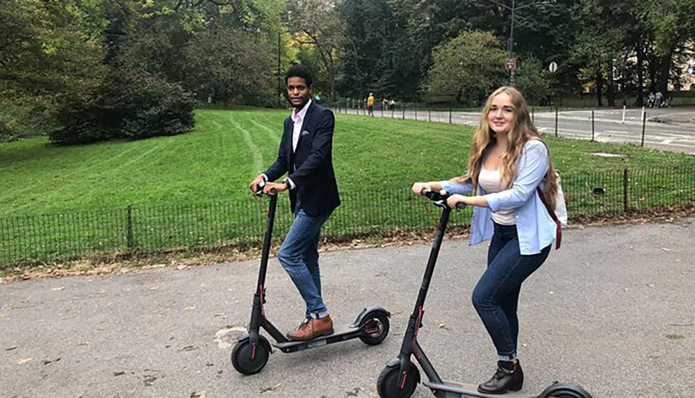 Two people are riding electric scooters along a path with greenery in the background