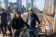 A smiling couple is posing with a bicycle on a crowded pedestrian walkway of the Brooklyn Bridge, with the Manhattan skyline in the background.
