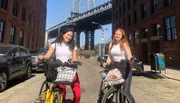 Two smiling individuals are standing with their bicycles on a sunny day with the backdrop of a bridge and brick buildings.