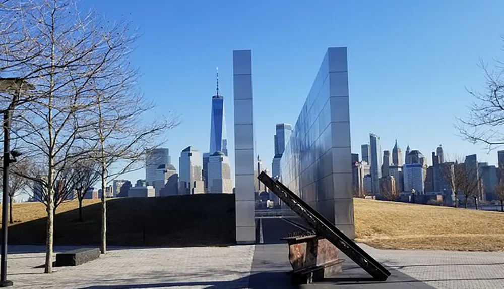 The image shows a modern memorial consisting of two tall reflective structures framing a pathway with a view of the Manhattan skyline and the One World Trade Center in the background