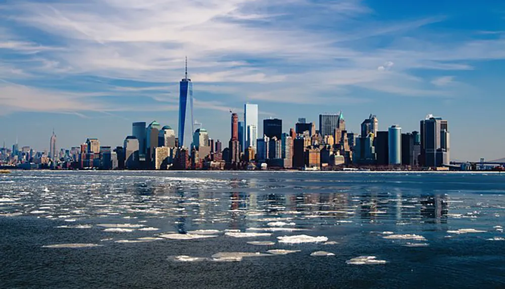 The image shows the skyline of Lower Manhattan New York City with ice floating on the water in the foreground under a blue sky with scattered clouds