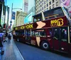 A double-decker Big Bus New York tour bus is navigating through a busy urban street with pedestrians and towering skyscrapers