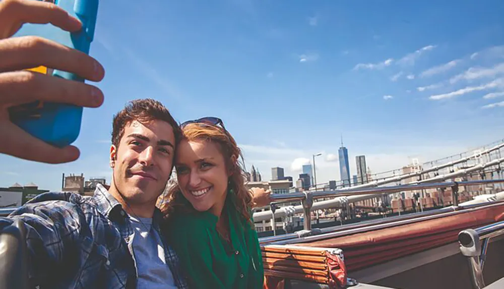 A young couple is taking a selfie with a city skyline in the background under a clear blue sky