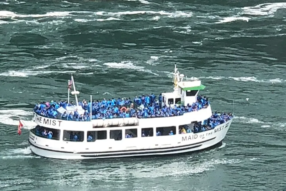 A crowded tour boat filled with passengers wearing blue ponchos is navigating a rippled body of water