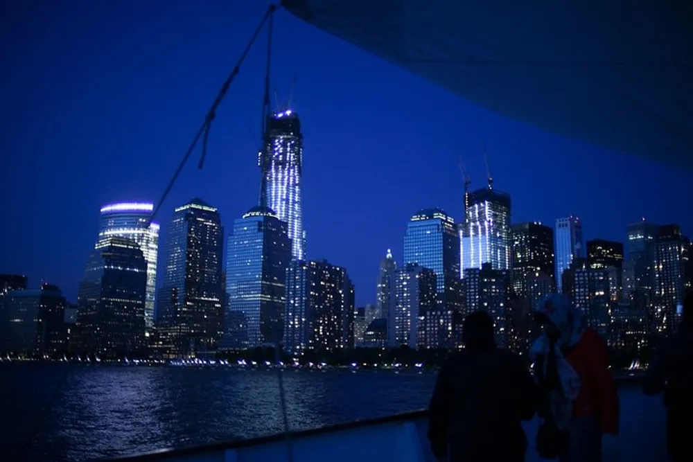 Passengers on a boat gaze at a city skyline illuminated against the evening sky