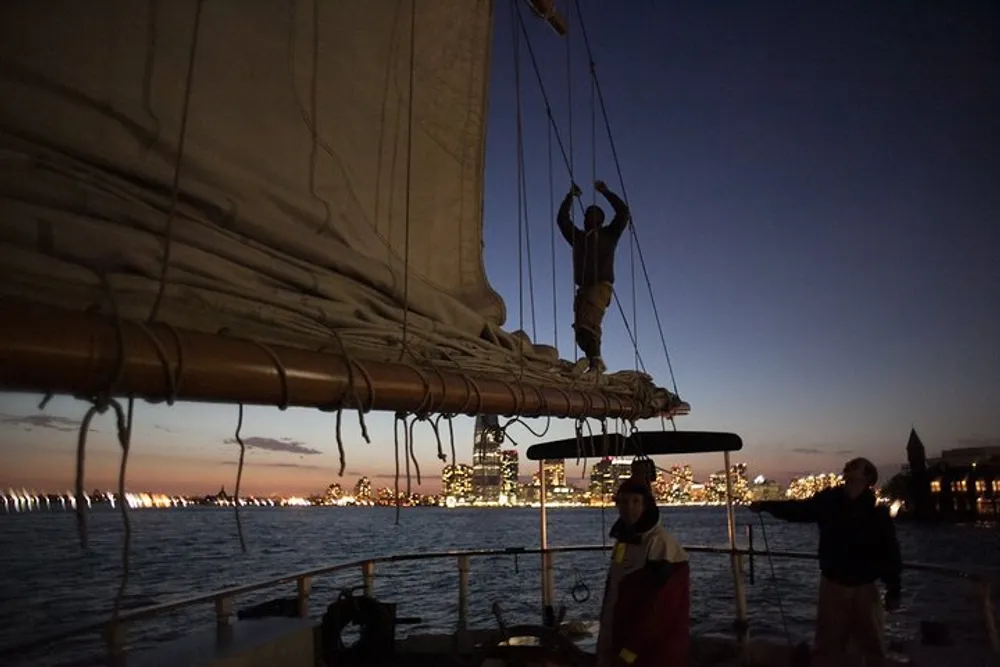 A person is attending to the sails on a boat at dusk with a city skyline illuminated in the background