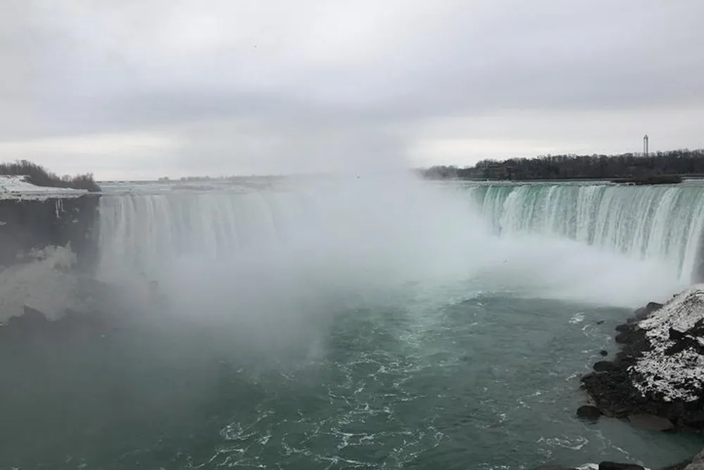 The image shows the misty and mighty Niagara Falls during a cloudy day with snow and ice along its banks