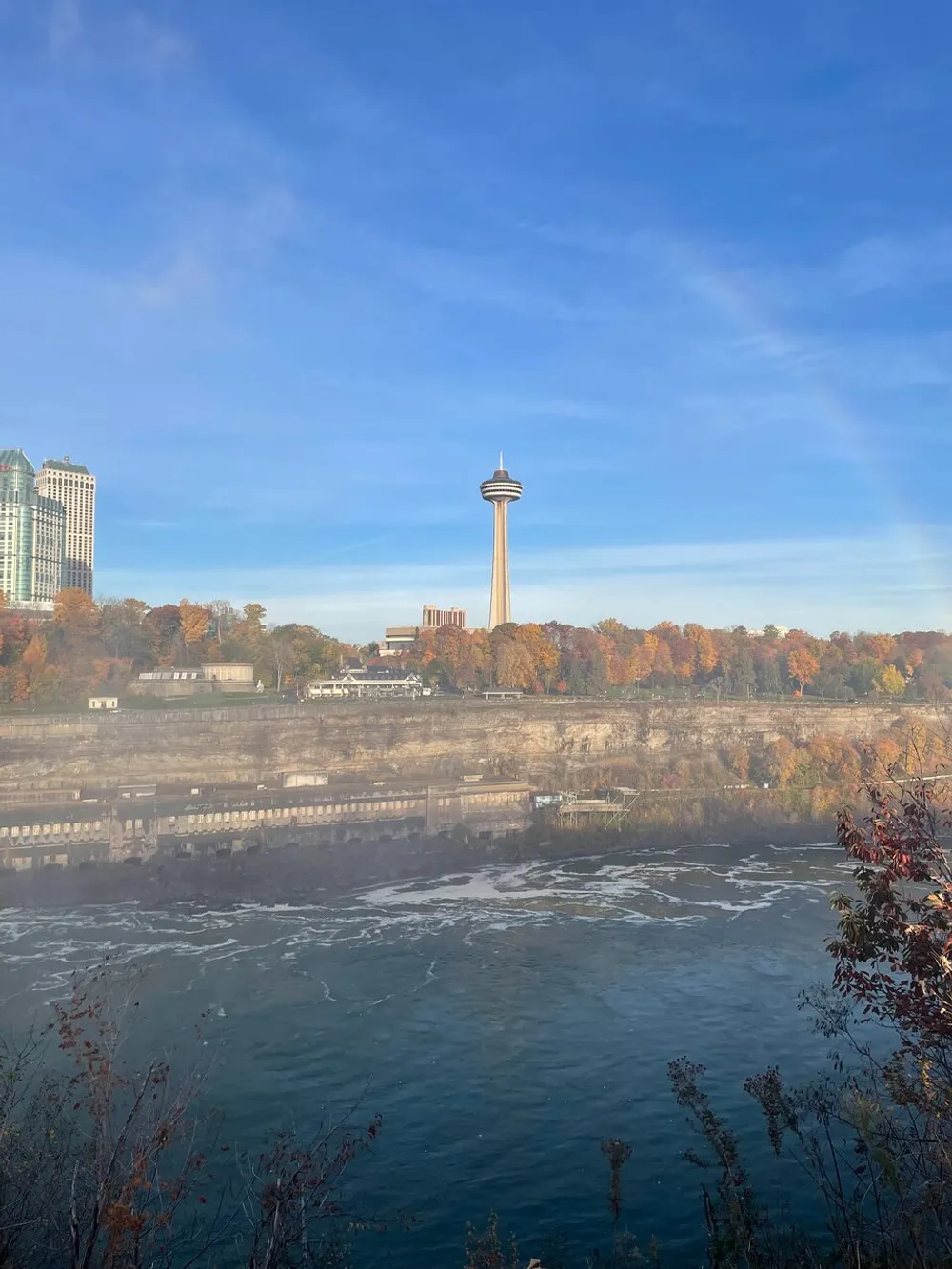 The image shows a scenic view with the Skylon Tower in the background a river in the foreground and autumn-colored trees highlighting a beautiful mix of natural and man-made landscapes