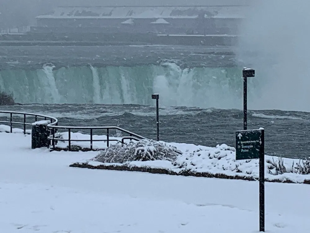 The image captures a snowy scene with a misty waterfall in the background likely Niagara Falls viewed from a railing-lined observation area with a direction sign partially visible