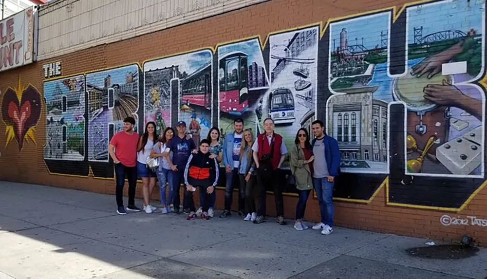 A group of people is posing for a photo in front of a colorful mural that spells out THE BRONX with various images representing urban life within each letter