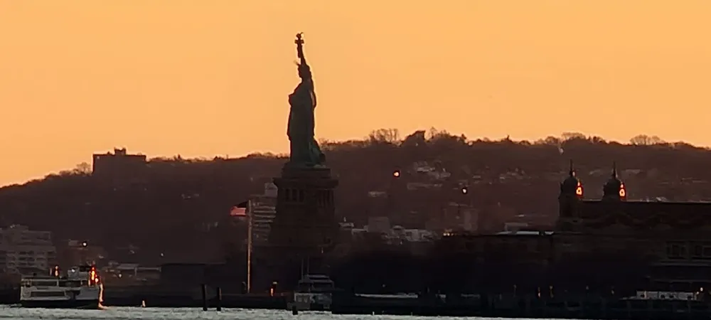 The image captures a silhouette of the Statue of Liberty against an orange-hued sunset sky