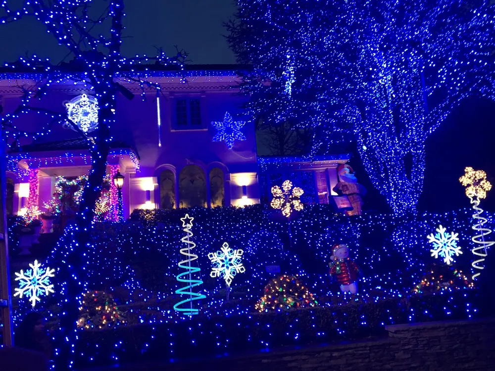 The image displays a house richly adorned with blue and white holiday lights and decorations including illuminated trees stars and snowflake motifs creating a vibrant and festive nighttime scene