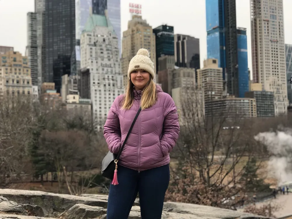 A person is smiling for the camera with a backdrop of skyscrapers that suggest an urban park setting possibly Central Park in New York City