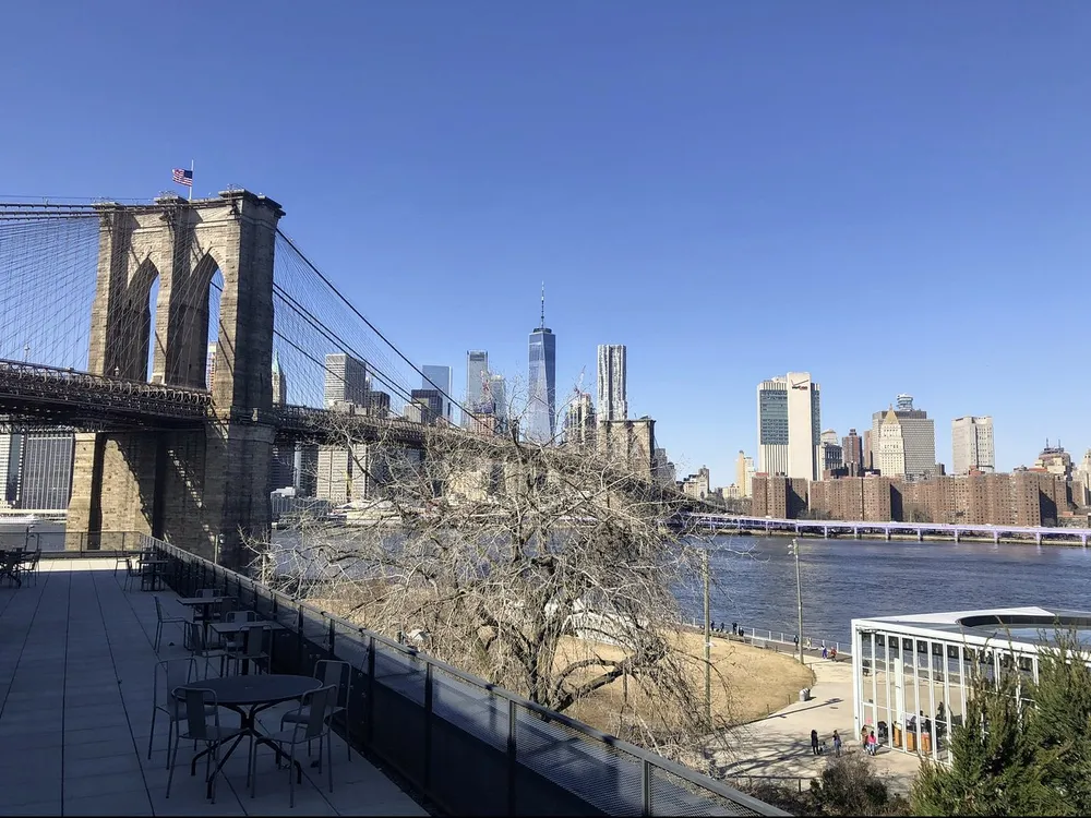 The photo features a clear view of the Brooklyn Bridge with the Lower Manhattan skyline in the background taken from an elevated vantage point that includes outdoor seating