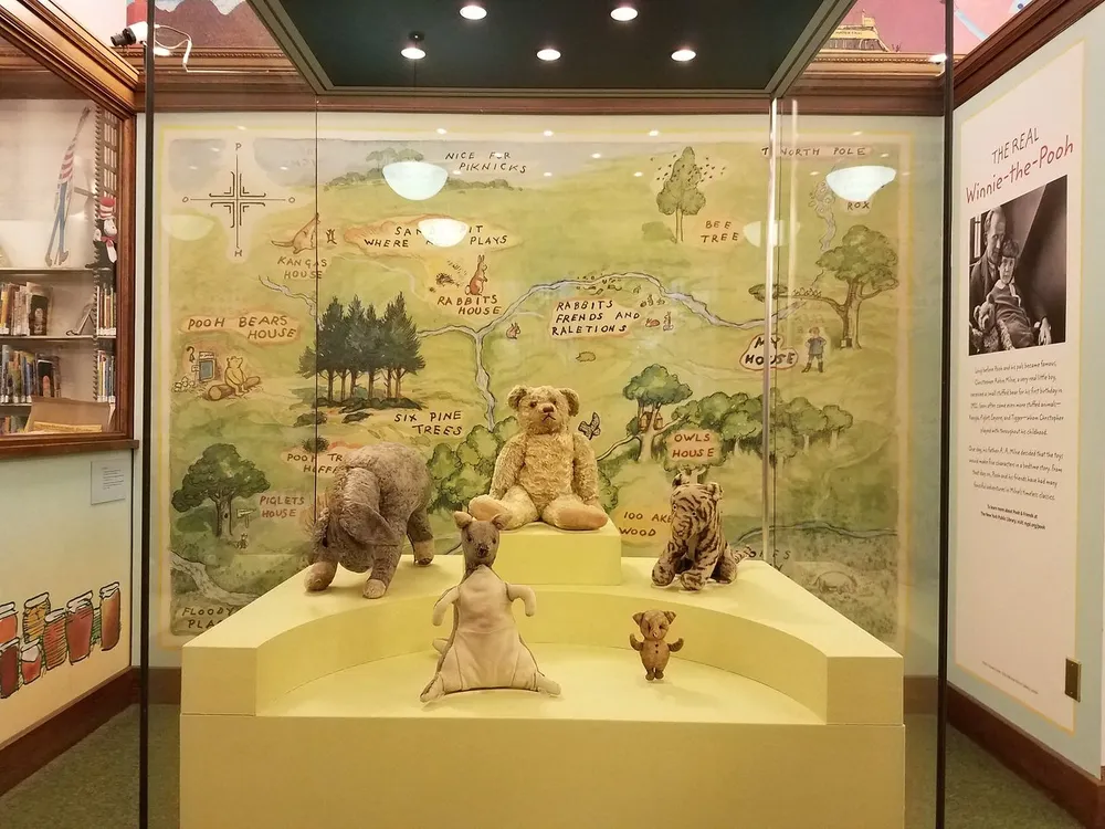 This image shows the original stuffed animals that inspired the characters in A A Milnes Winnie-the-Pooh stories displayed in a glass case with a backdrop illustrating the fictional Hundred Acre Wood