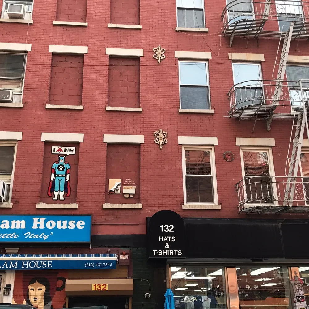 A red brick building with eclectic decorations including a pixel-art style figure by a window and a fire escape on the right houses businesses including one that sells hats and t-shirts