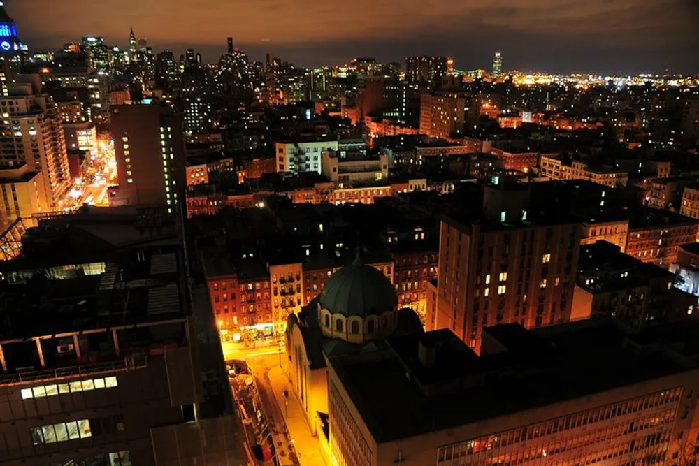 The image captures a vibrant nighttime cityscape with illuminated streets and buildings a prominent domed structure in the foreground and a backdrop of densely packed urban towers under a dark sky