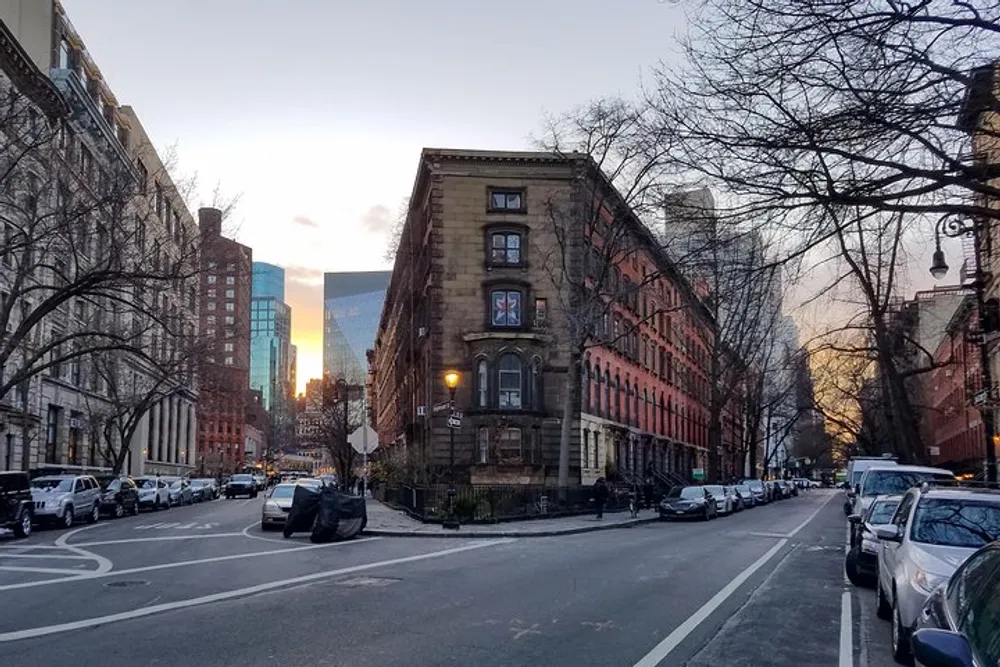 The image shows a city street at dusk with a prominent wedge-shaped building parked cars and bare trees against a backdrop of sunlit modern skyscrapers