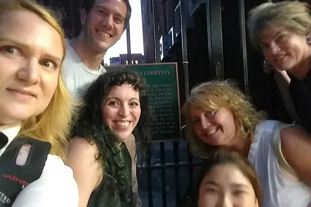 A group of smiling people are taking a selfie together near what appears to be an entrance to a cemetery as indicated by a sign