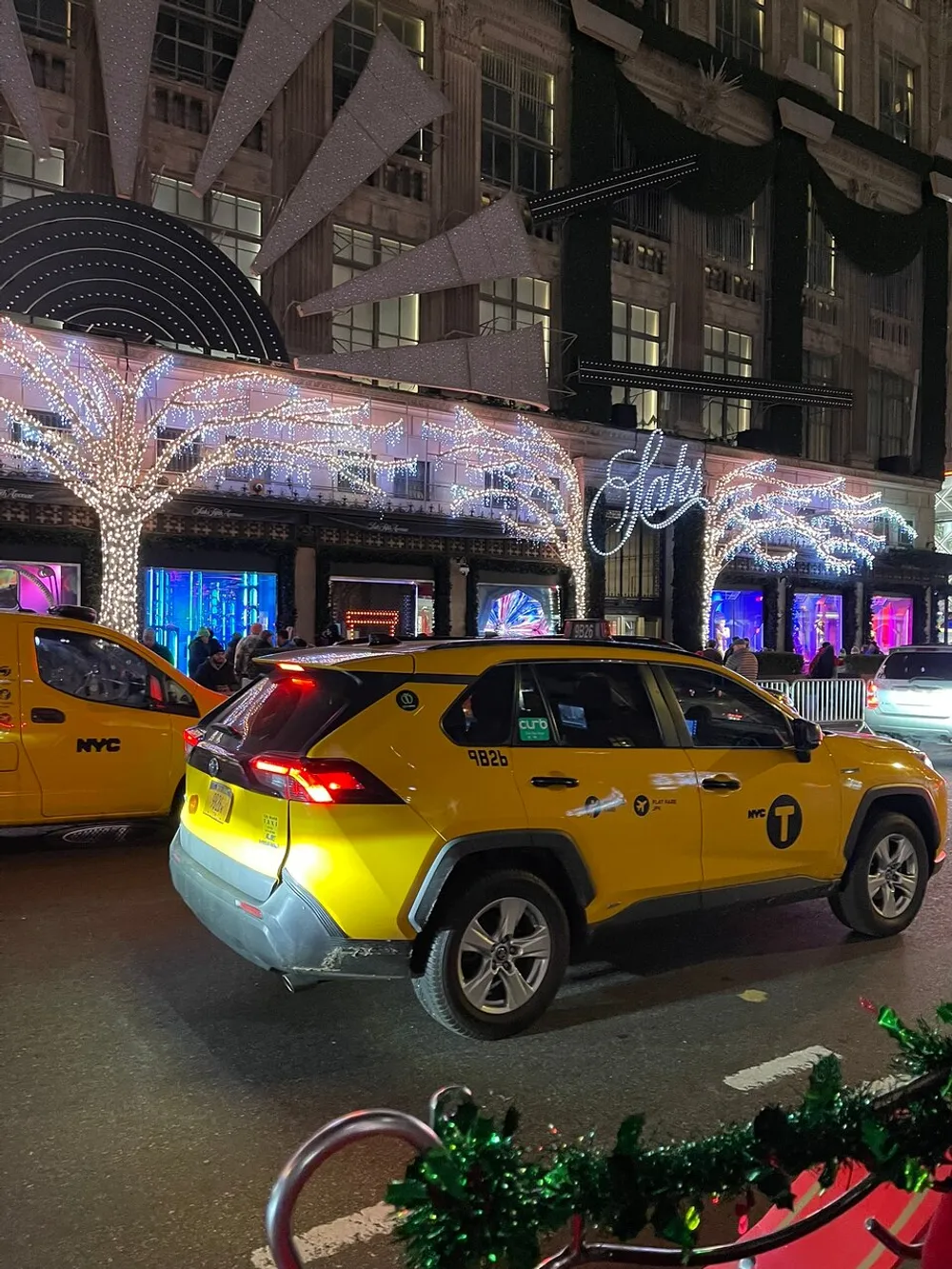 An iconic yellow NYC taxi is passing by a festively decorated building with large illuminated stars and glowing trees at night