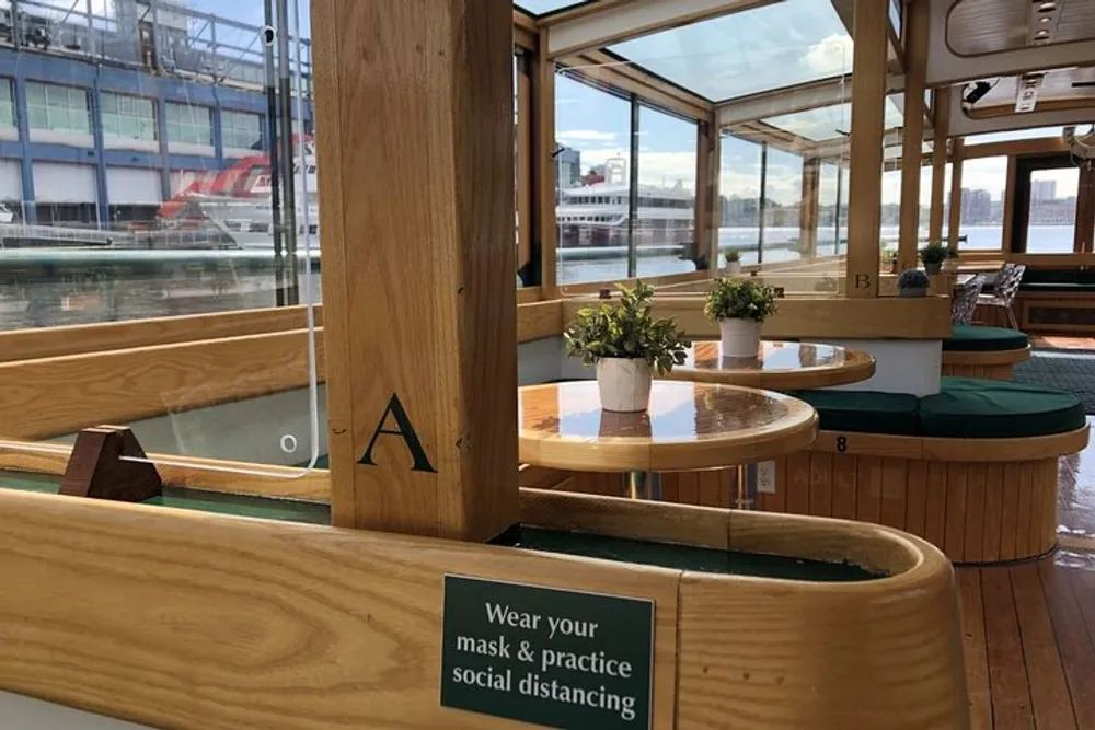 The image shows an interior section of a boat with wooden seating and tables potted plants and a sign advising to wear a mask and practice social distancing indicative of health precautions during the COVID-19 pandemic