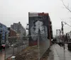 An urban street scene features a large mural of a person with their chin resting on their hands painted on the side of a building with the Williamsburg Bridge in the background