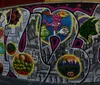 An urban street scene features a large mural of a person with their chin resting on their hands painted on the side of a building with the Williamsburg Bridge in the background