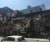 An urban street scene features a large mural of a person with their chin resting on their hands painted on the side of a building with the Williamsburg Bridge in the background