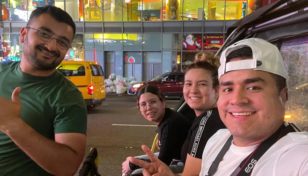 Four individuals are posing for a selfie at night with a vibrant city street in the background featuring a yellow cab and neon-lit storefronts