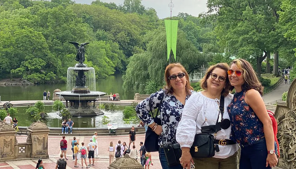 Three individuals are posing for a photo in front of the Bethesda Fountain in Central Park New York City with many visitors enjoying the surroundings