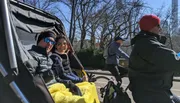 Two people are enjoying a ride in a pedicab on a sunny day with cyclists and bare trees in the background.