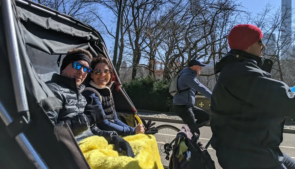 Two people are enjoying a ride in a pedicab on a sunny day with cyclists and bare trees in the background