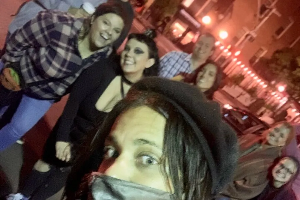 A group of people are smiling and posing for a selfie at night with one person in the foreground wearing a mask and a hat