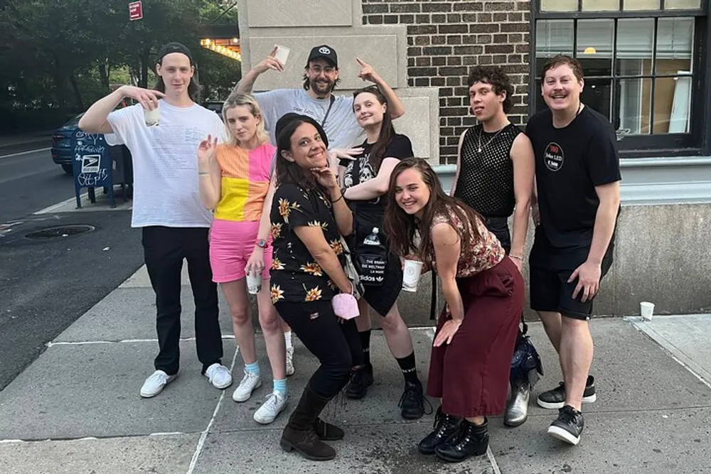 A group of eight individuals is posing playfully for a photo on a sidewalk with some making gestures towards the camera and others smiling or crouching creating a casual and jovial atmosphere