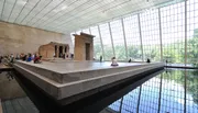The image shows an ancient Egyptian temple structure exhibited within a large, light-filled room featuring a glass wall and a reflecting pool, with visitors observing and resting around the space.