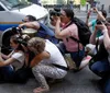 A group of photographers is crouched down aiming their cameras towards something of interest out of the frame near a police vehicle on the street