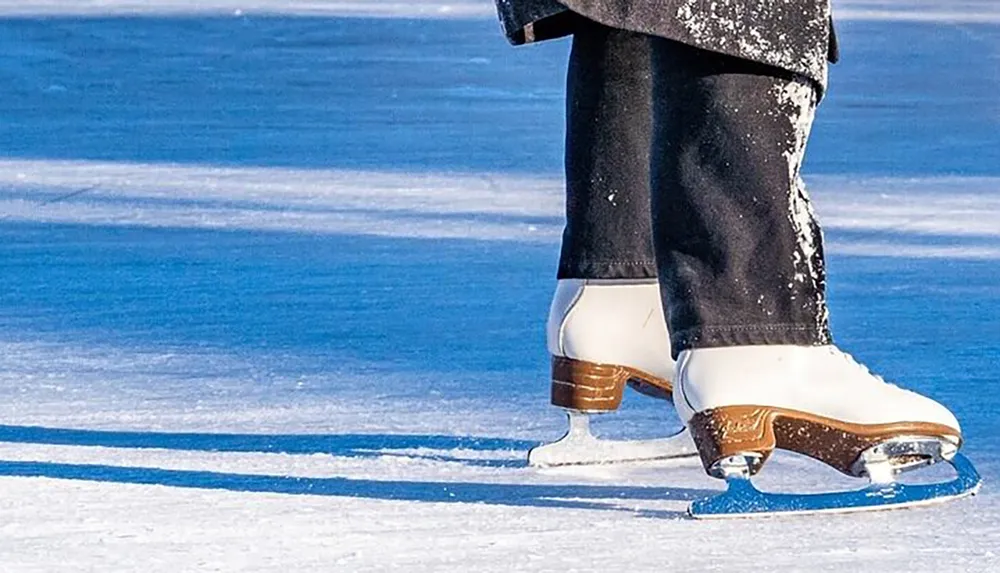 A person is shown from the knee down wearing white figure skates on an ice rink